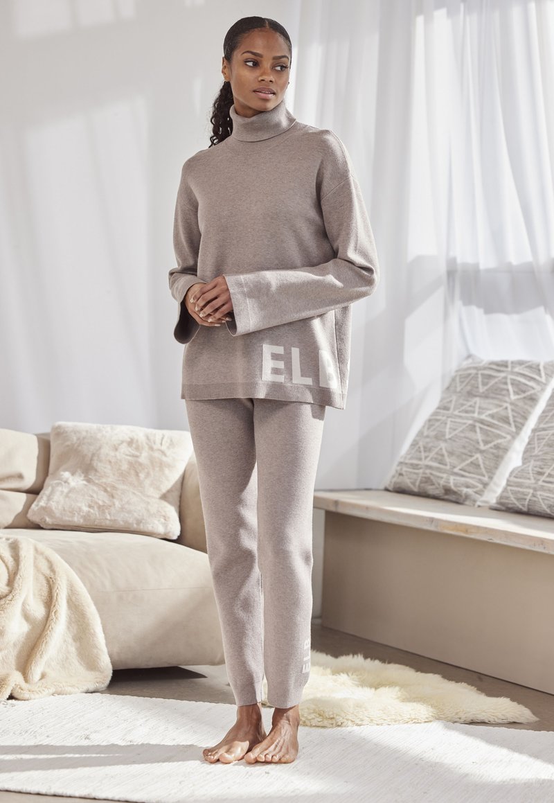 Beige Strick-Turtleneck-Pullover mit weiten Ärmeln und Logodetail am Saum, kombiniert mit passenden, eng anliegenden Hosen, beide aus einem weichen Stoff.