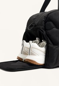 Sac à dos noir avec une poche avant ouverte contenant une paire de chaussures de sport blanches avec des semelles marron à l'intérieur.
