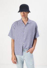 Camisa de manga corta abotonada en un patrón geométrico azul y blanco. Tejido suave, corte relajado. Combinada con un sombrero de cubo azul marino y jeans claros.