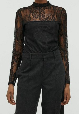 Femme portant un haut noir en dentelle florale à manches longues avec un col montant et un pantalon gris foncé tailleur, sur un fond uni.