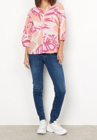 Blusa colorata e comoda con scollatura a V e dettagli a bottoni, caratterizzata da motivi astratti di foglie rosa e pesco. Indossata con jeans skinny blu e sneaker bianche.