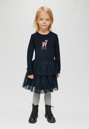 Jeune fille aux cheveux blonds portant une robe bleu marine avec une jupe en tulle superposée, un motif de cerf rose, des collants gris et des bottes à lacets noires se tenant sur un fond blanc.