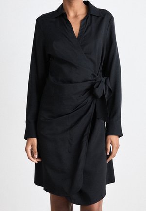 Kobieta ubrana w czarną, długorękawową sukienkę kopertową z kołnierzykiem i wiązaniem z boku, stojąca na jednolitym jasnym tle.