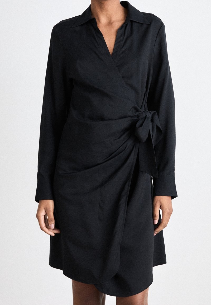 Kobieta ubrana w czarną, długorękawową sukienkę kopertową z kołnierzykiem i wiązaniem z boku, stojąca na jednolitym jasnym tle.