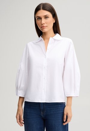 Femme aux cheveux bruns raides portant un chemisier blanc boutonné avec des manches trois-quarts bouffantes et un jean bleu sur un fond uni.