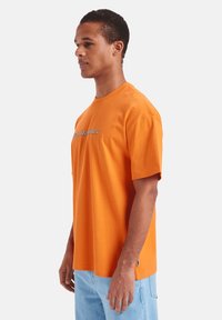 Oranžové bavlněné tričko s krátkými rukávy, které na přední straně zdobí logo v kontrastních barvách a má standardní střih.
