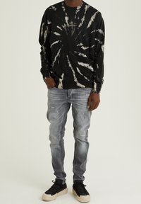 Sudadera negra con diseño de tie-dye y patrones en espiral blancos, combinada con jeans claros desgastados y zapatillas negras con acentos crema.