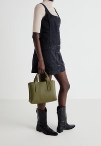 Robe salopette en denim noir avec des boutons à l'avant et deux poches latérales, associée à des bottes de cowboy noires et un sac à main en olive texturé.