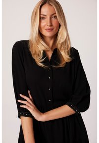 Czarna sukienka na guziki z rękawami o długości trzech czwartych. Posiada metaliczne guziki i teksturowane akcenty na końcach rękawów. Luźny krój.