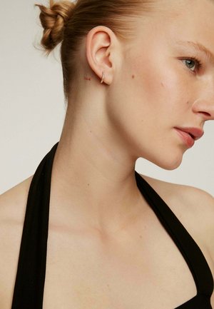 Pendiente de aro de oro con un pavé de pequeñas piedras transparentes. La modelo lleva una blusa negra de cuello halter, tiene piel clara y un peinado de moño sutil.