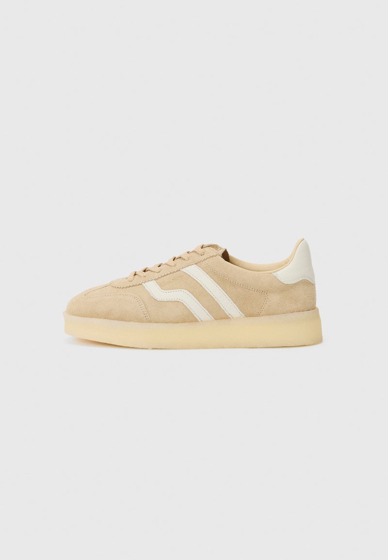 Sneaker in suede beige con strisce laterali bianche, punta arrotondata, chiusura con lacci e suola testurizzata. Design minimalista e colori neutri.
