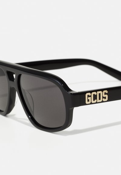 Black sunglasses with dark lenses, featuring a matte finish and gold "GCDS" logo on the temple. Rectangular shape and contemporary design.