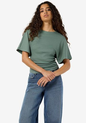 Kvinde med langt krøllet hår iført en grøn kortærmet top og blå jeans, står med hænderne løst sammenlagt foran.