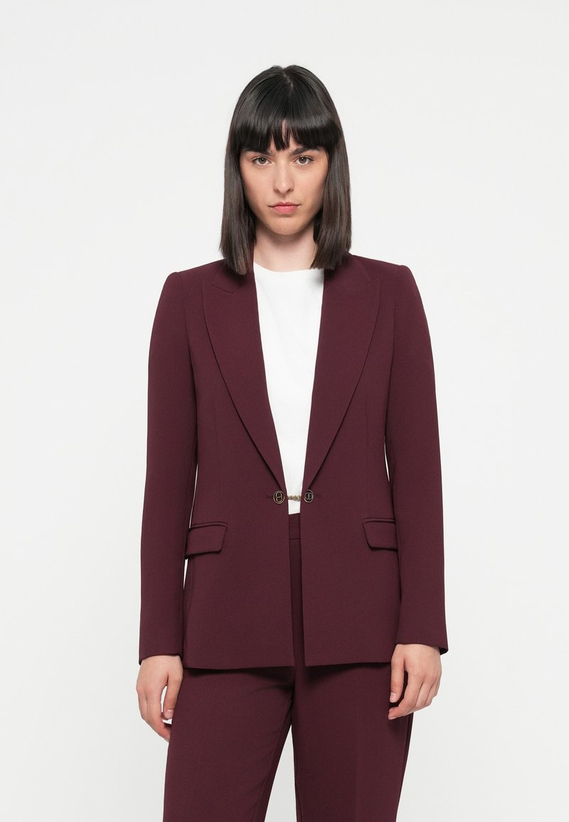 Burgundy tailored blazer with a satin finish, notch lapels, and gold hardware. Features two front pockets and a single button closure.