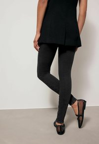 Mustat legginsit, joissa on sileä pinta, tiukka muotoilu ja keskikorkea vyötärö. Yhdistettynä litteisiin, mustiin, verkkomaisiin kenkiin, joissa on hihnatiedot.