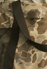 Sac à motif camouflage en beige et gris atténué avec une poche en maille. Présente un logo et des poignées robustes en marron. Fabriqué en tissu durable.