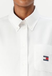Hvit skjorte med krage, med en brystlomme og broderte "TOMMY JEANS" logo i rød, hvit og marineblå.
