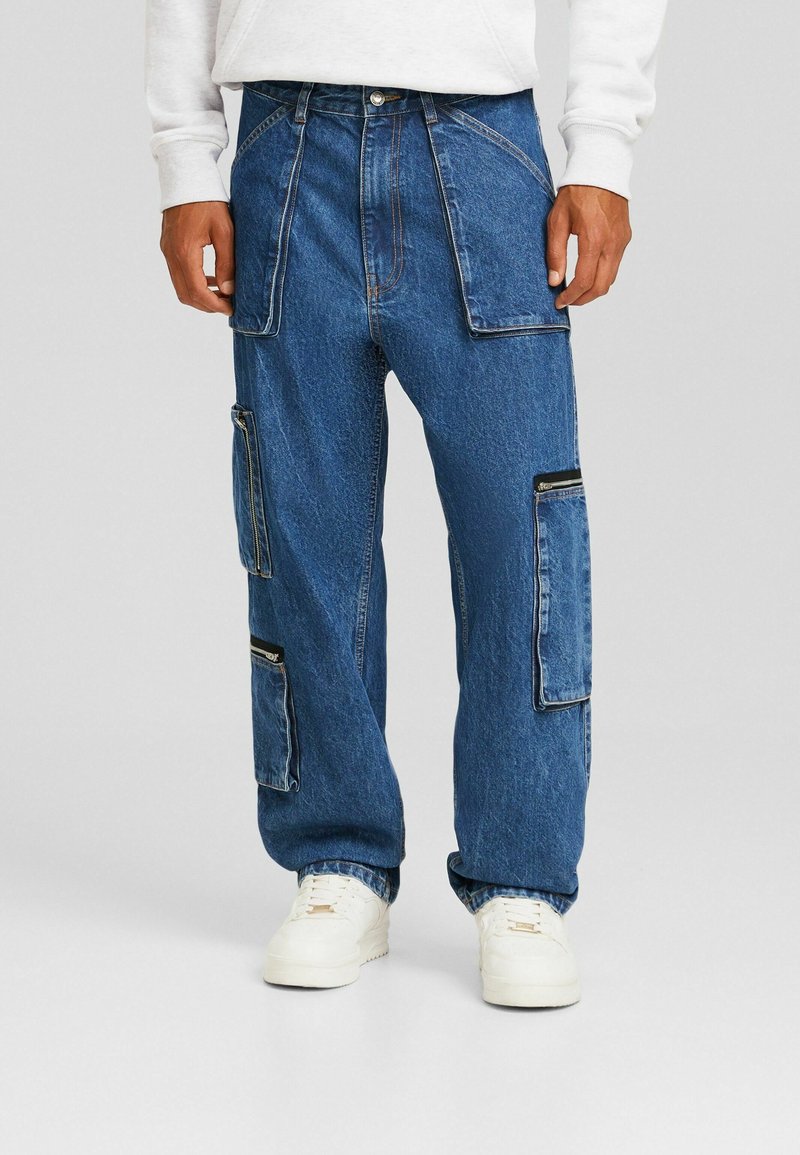 Pantalones cargo de mezclilla azul con múltiples bolsillos, corte holgado, costuras visibles y detalles con cremalleras. Combinados con zapatillas blancas.