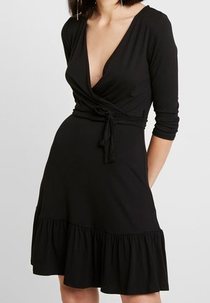 Femme portant une robe portefeuille noire longueur genoux avec manches 3/4, un profond décolleté en V, une taille nouée et un ourlet à volants sur un fond uni.