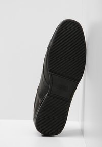 Chaussure de sport noire avec une tige texturée, une semelle en caoutchouc à rainures horizontales, et un logo contrastant sur le côté pour l'adhérence et le style.