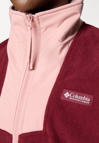 Burgunderfarget og rosa fleecejakke med høy krage, frontglidelås og Columbia-logo. Har en glatt rosa overdel og en teksturert burgunder underdel.