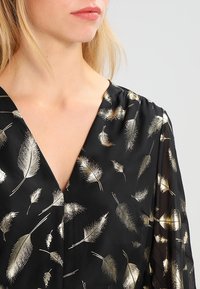 Blusa nera con stampa di piume dorate. Presenta un profondo scollo a V e spalle arricciate. Il tessuto ha una texture liscia e leggermente lucida.
