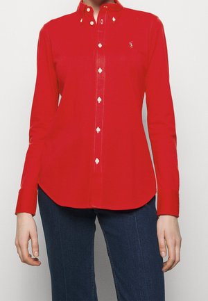 Mujer con una camisa roja ajustada abotonada de manga larga y vaqueros azul oscuro, de pie frente a un fondo liso.