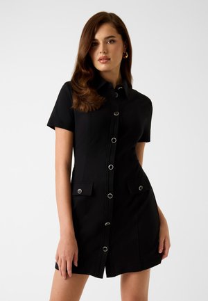 Schwarzes Kurzarmkleid mit durchgehender Knopfleiste, silbernen Beschlägen und zwei Taschen vorne. Das Kleid ist figurnah geschnitten und hat eine glatte Textur.