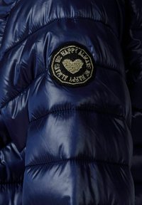 Marinblå pufferjacka med en glänsande yta, med en rund svart märkning med broderad text och hjärtmotiv på den övre ärmen.