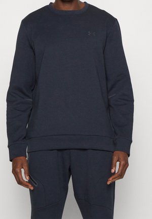 Mężczyzna ubrany w granatową bluzę z długim rękawem marki Under Armour i pasujące spodnie na jednolitym jasnym tle.