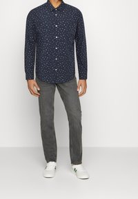 Marineblauw overhemd met kleine, multicolor patronen, lange mouwen en een afgeronde zoom. Gecombineerd met grijze jeans en witte sneakers.