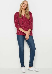 Blusa leopardata rossa e nera con scollo a V e maniche a tre quarti, abbinata a jeans skinny blu e sneaker bianche.