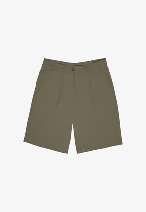 Mann iført beige shorts, en flerfarget kortermet skjorte, hvite sokker og sølvfargede treningssko som står med hendene i lommene.