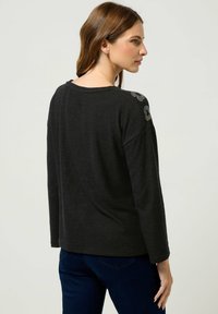 Schwarzes Langarmtop mit entspannter Passform, verziert mit Schulterdetails und einer weichen Textur. Der Rücken hat einen abgerundeten Ausschnitt.