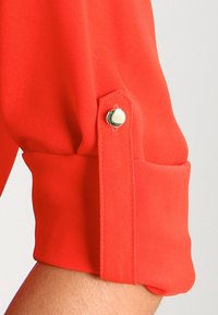 Röd blus med detalj på ärmarna och en snug passform, med en manschett som fästs med en silverknapp och en slät, lätt tygtextur.