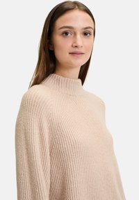 Beige gestrickter Rollkragenpullover mit strukturiertem Rippmuster, der einen hohen Kragen und Raglanärmel für eine lässige Passform hat.