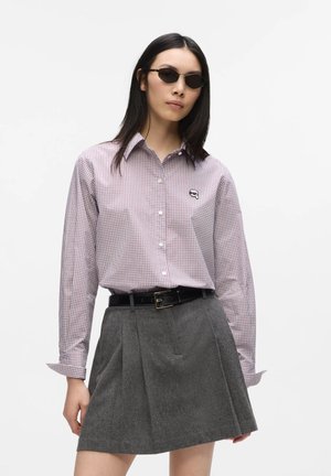 Femme portant des lunettes de soleil, une chemise à carreaux rose boutonnée rentrée dans une jupe grise plissée avec une ceinture noire, debout devant un fond blanc.