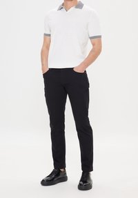 Bílá polokošile s límečkem a manžetami s černými pruhy, v kombinaci s černými kalhotami úzkého střihu a černými teniskami. Hladká tkanina, minimalistický design.