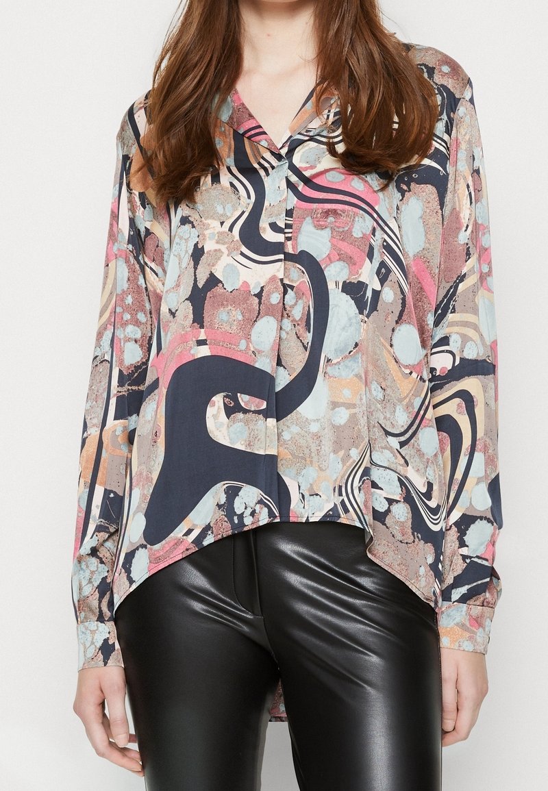 Femme portant un chemisier coloré à motif abstrait avec des manches longues et un pantalon en cuir noir, debout devant un fond uni.