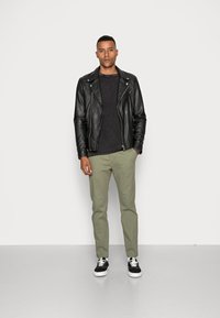 Schwarze Leder-Bikerjacke über einem dunklen Pullover, kombiniert mit olivgrünen Chinos und schwarzen Sneakers mit weißen Sohlen. Schlichte, figurbetonte Silhouette.