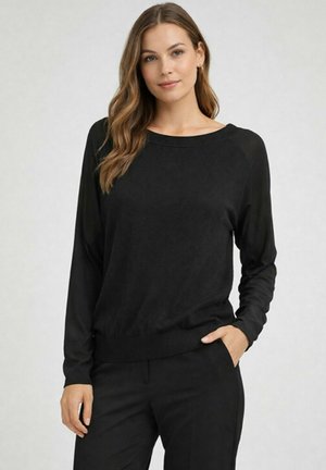 Femme aux cheveux châtain clair portant un pull noir à manches longues et un pantalon noir, debout avec une main dans la poche sur fond gris.