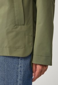 Chaqueta verde oliva con un acabado suave y texturizado. Presenta un bolsillo lateral y un corte relajado, combinada con pantalones de mezclilla.