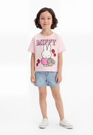 Enfant aux cheveux courts et foncés portant un t-shirt rose Miffy avec des fraises et un short en jean, debout sur un fond blanc.