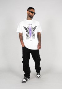 White graphic t-shirt featuring angel illustrations, purple accent, black text, paired with black cargo pants and black and white sneakers.