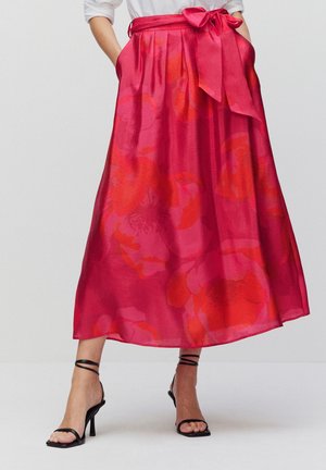 Femme portant une jupe midi rouge à fleurs avec une grosse ceinture nouée en nœud, associée à des sandales à talons hauts noires à brides et un haut blanc.
