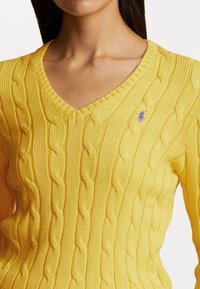 Gul stickad tröja med V-ringning, strukturerad kabeldesign och en liten broderad logotyp i blått på bröstet.