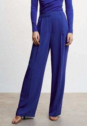 Model draagt een hooggetailleerde, wijd uitlopende broek in kobaltblauw met een bijpassend shirt met lange mouwen en tan open-toe sandalen met hak.