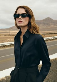 Czarna satynowa koszula z kołnierzykiem, w zestawie z czarnymi spodniami o wysokim stanie. Dodatki to ciemne okulary przeciwsłoneczne, na neutralnym tle.