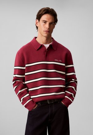Junger Mann trägt ein rotes Poloshirt mit weißen und schwarzen horizontalen Streifen und dunkle Hose, steht mit den Händen in den Taschen vor grauem Hintergrund.