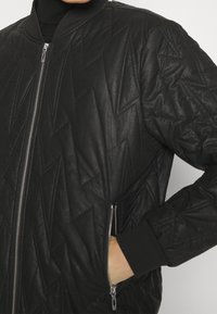 Veste bomber matelassée noire avec un motif géométrique, fermeture éclair et poignets côtelés. Fabriquée en matériau imitant le cuir avec des poches latérales.
